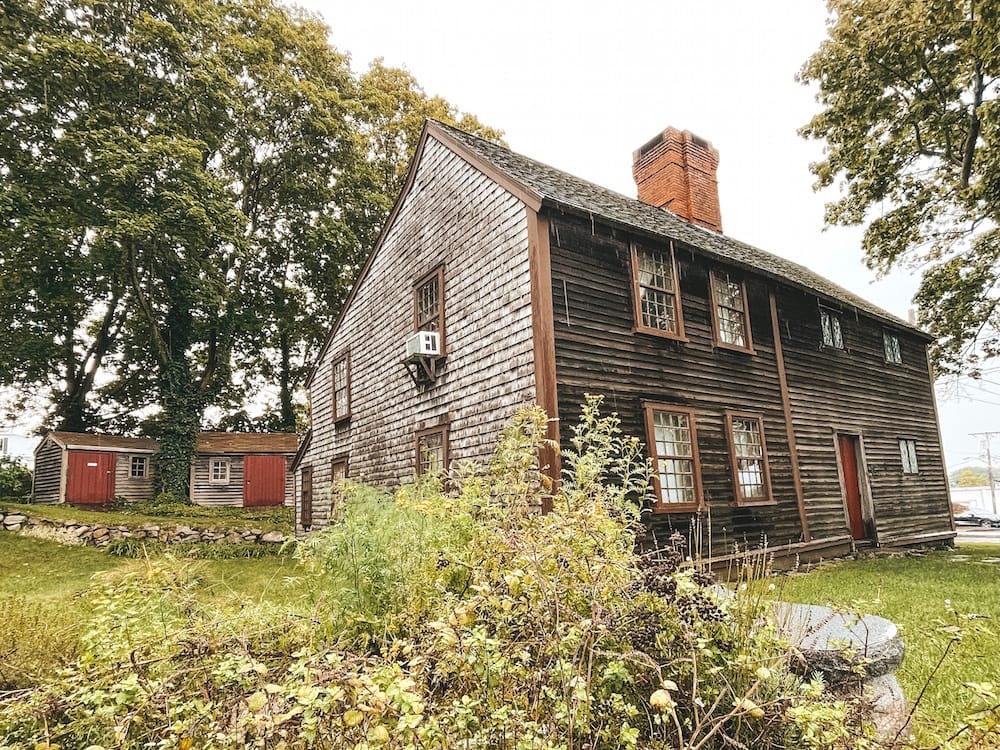 An old wooden house in Plymouth, MA, surrounded by greenery with red doors and a brick chimney.