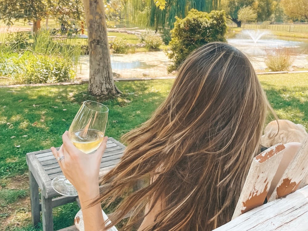A woman with brown hair sitting and drinking a glass of white wine in a green winery overlooking a lake.