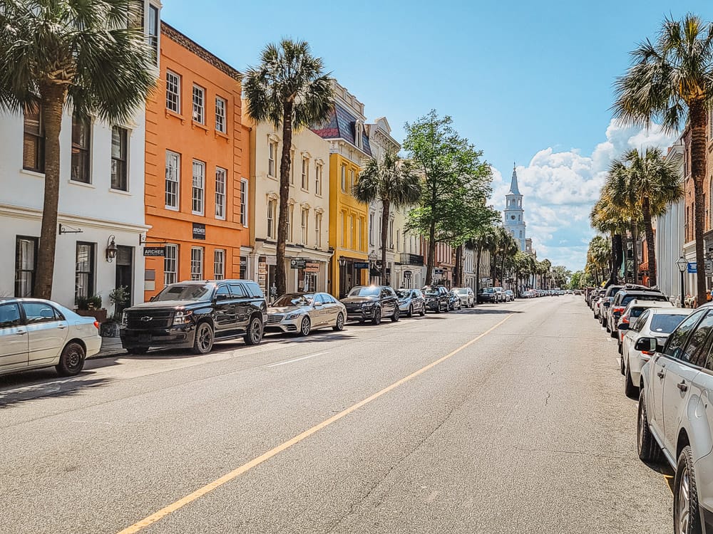 A street lined with cars and colorful buildings and a church in the background in Charleston, SC