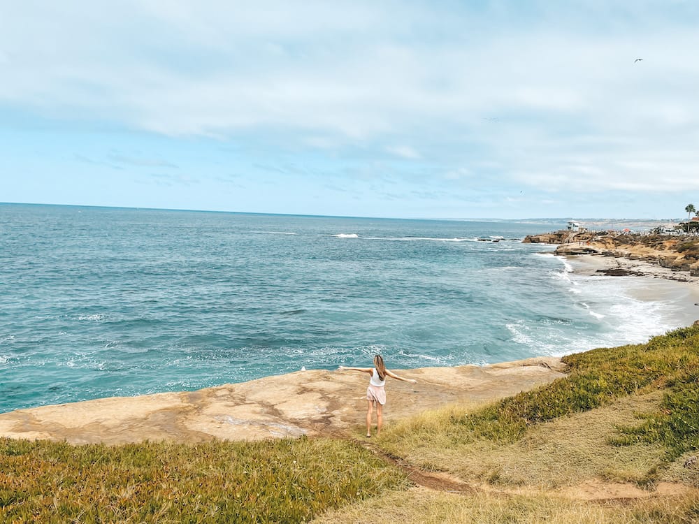 A woman standing on a sandstone cliff holding out her arms as the blue waves crash onto the shore in San Diego