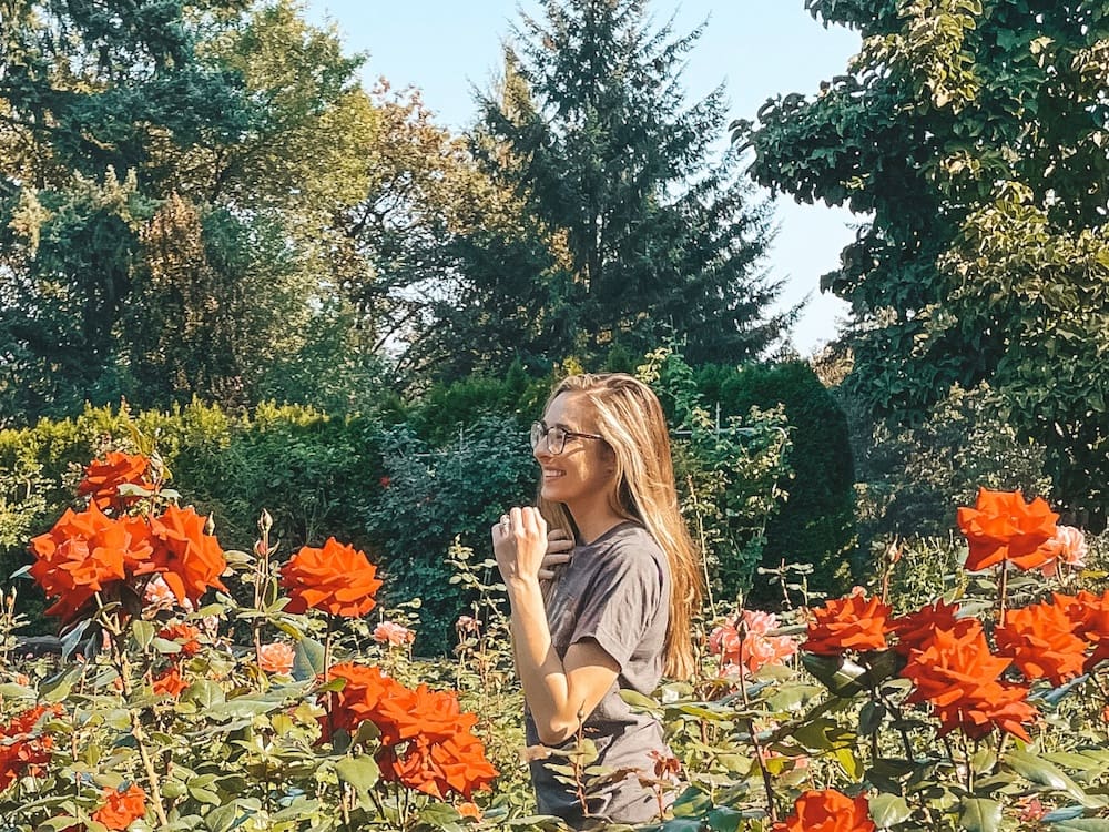 A woman in a gray shirt with brown hair and glasses standing in a red rose garden in Portland, Oregon, in October.