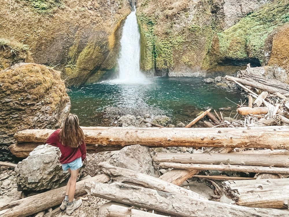 A woman in a red sweatshirt and denim shorts standing in front of a waterfall and blue pool of water in Portland - one of the best places to visit in the USA in August.