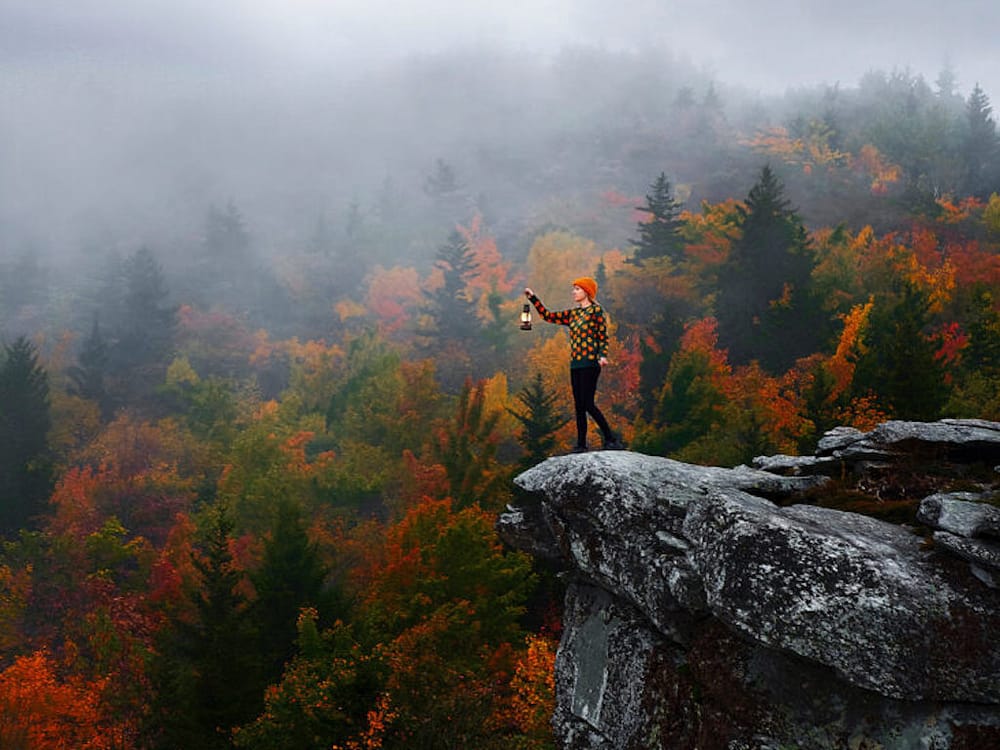 A woman standing on the ledge of a rock in Asheville, NC, with fall foliage in the background.
