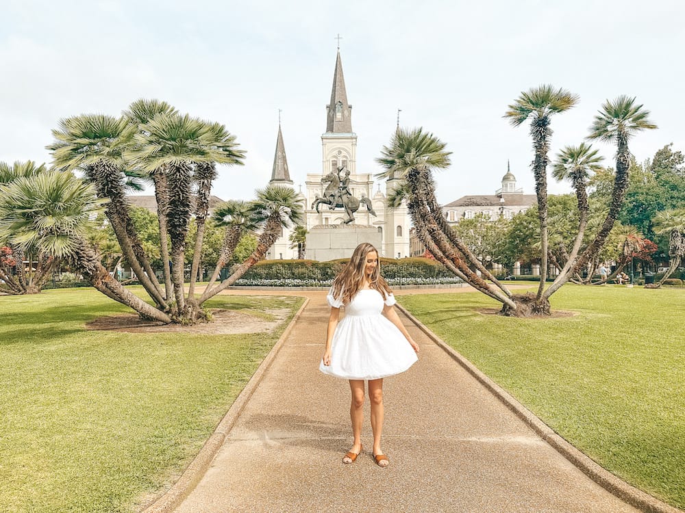 A woman in a white dress standing in Jackson Square in New Orleans, a park with greenery and a white church and a statue of a man riding a horse.
