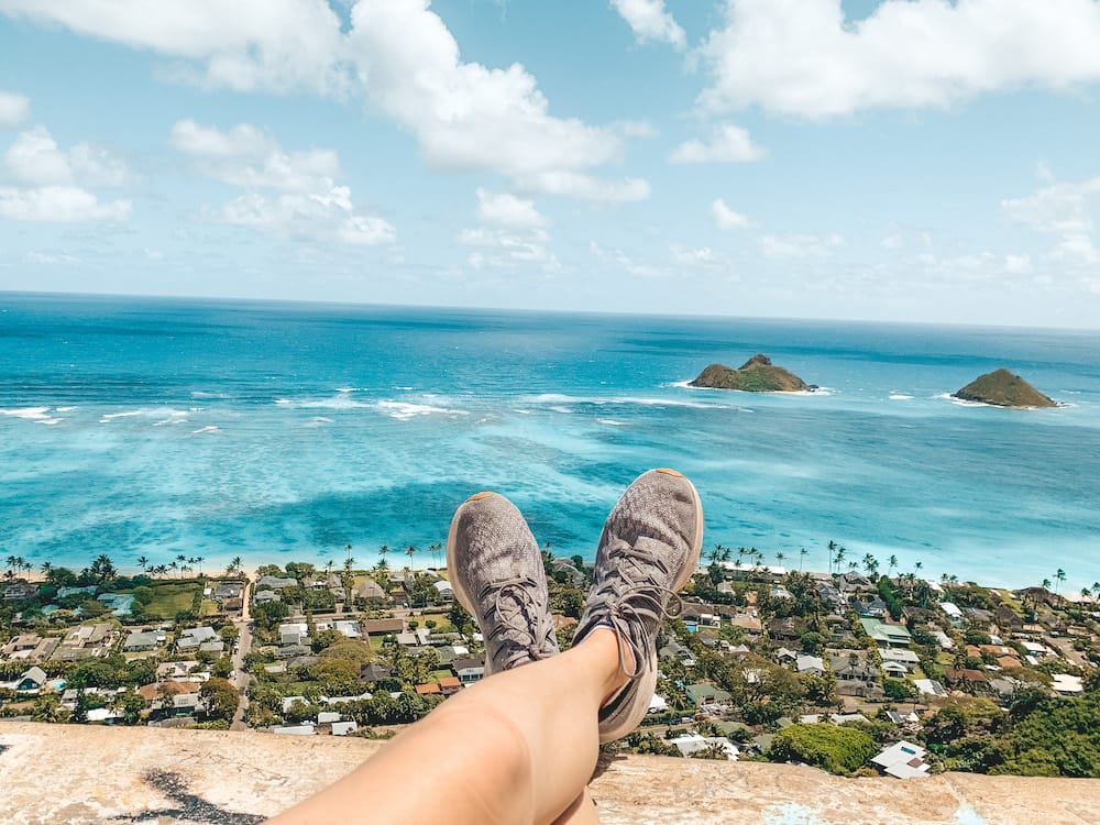 A pair of grey tennis shoes dangling over the edge of the pillbox on the Lanikai Pillbox Hike with the blue ocean, neighborhood, and green islands in the background.