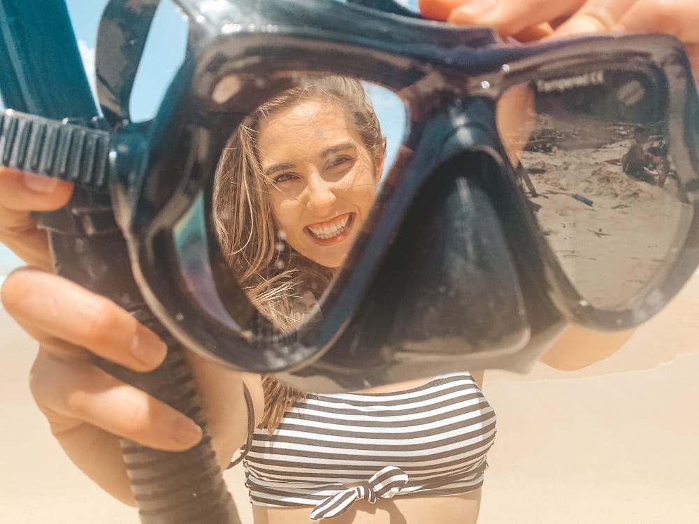 A girl holding a pair of snorkeling goggles up to the camera and smiling through the lens of one side wearing a black and white striped swimsuit.