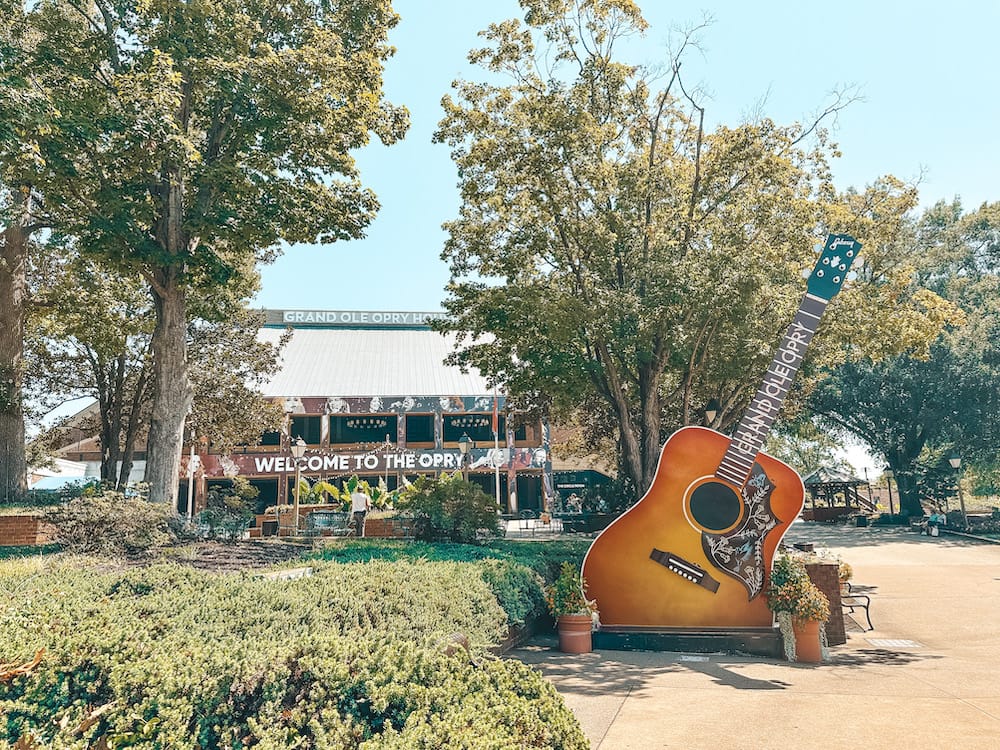 A snapshot of the giant orange guitar in front of the Grand Ole Opry building in Nashville, Tennessee. The photo is framed by greenery.