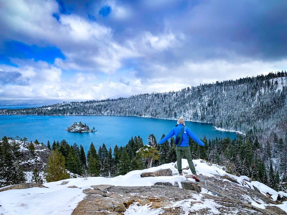 A woman wearing a blue puffer jacket standing in front of a stunning winter landscape featuring a blue alpine lake, snow-covered pine trees, and snow-covered rocks.