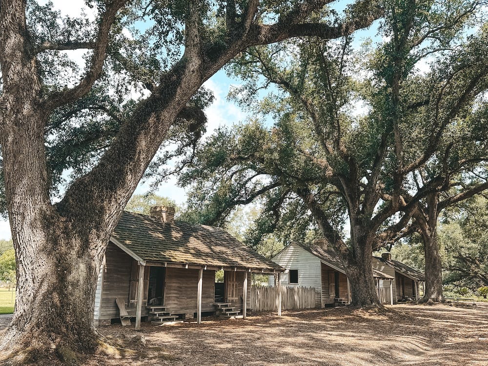 The Slavery at Oak Alley Exhibit, featuring two wooden buildings with oak trees providing them with shade.