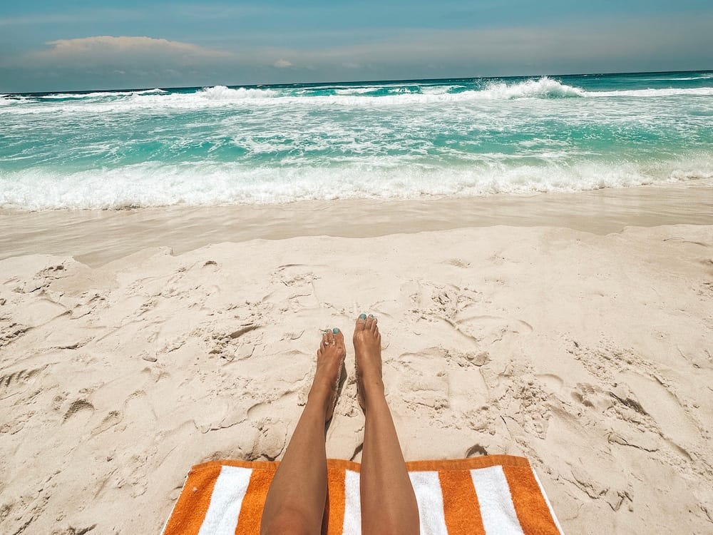 Two tanned legs on a orange and white beach towel on a sandy beach with turquoise waves lapping onto the shore
