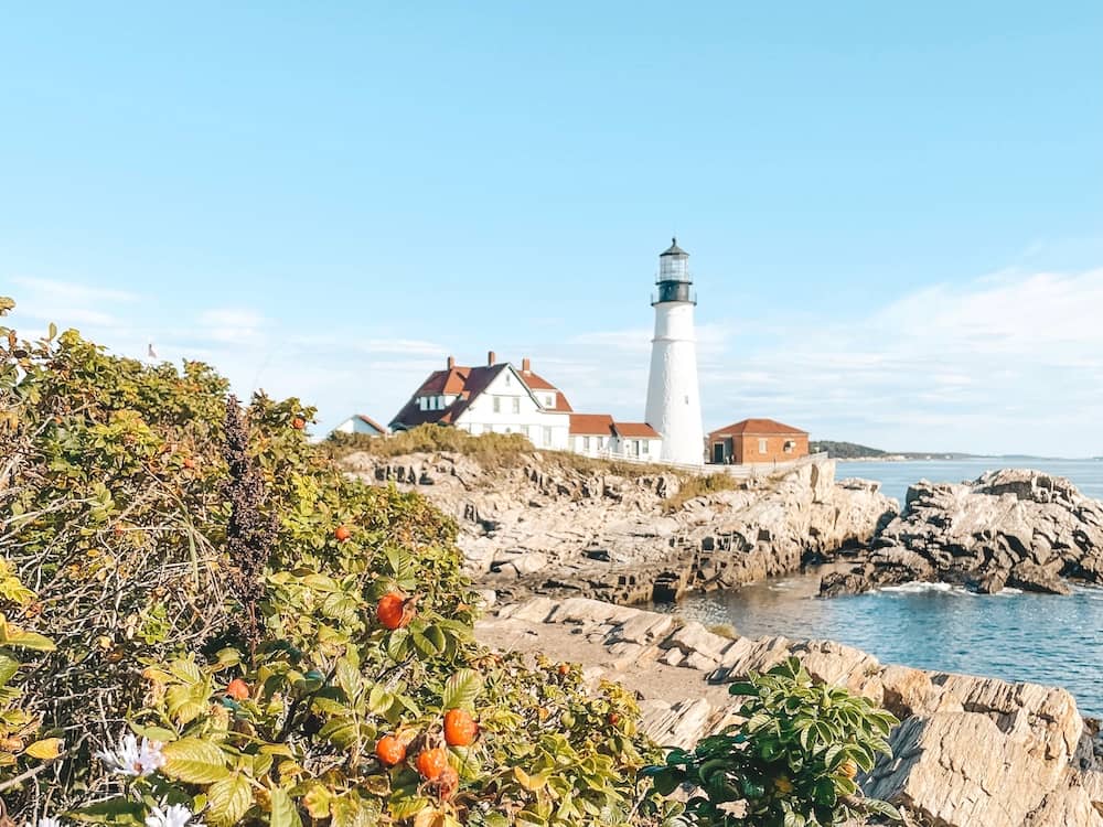 A white lighthouse surrounded by orange-red buildings on a rocky coastline in Maine, with greenery in the foreground.
