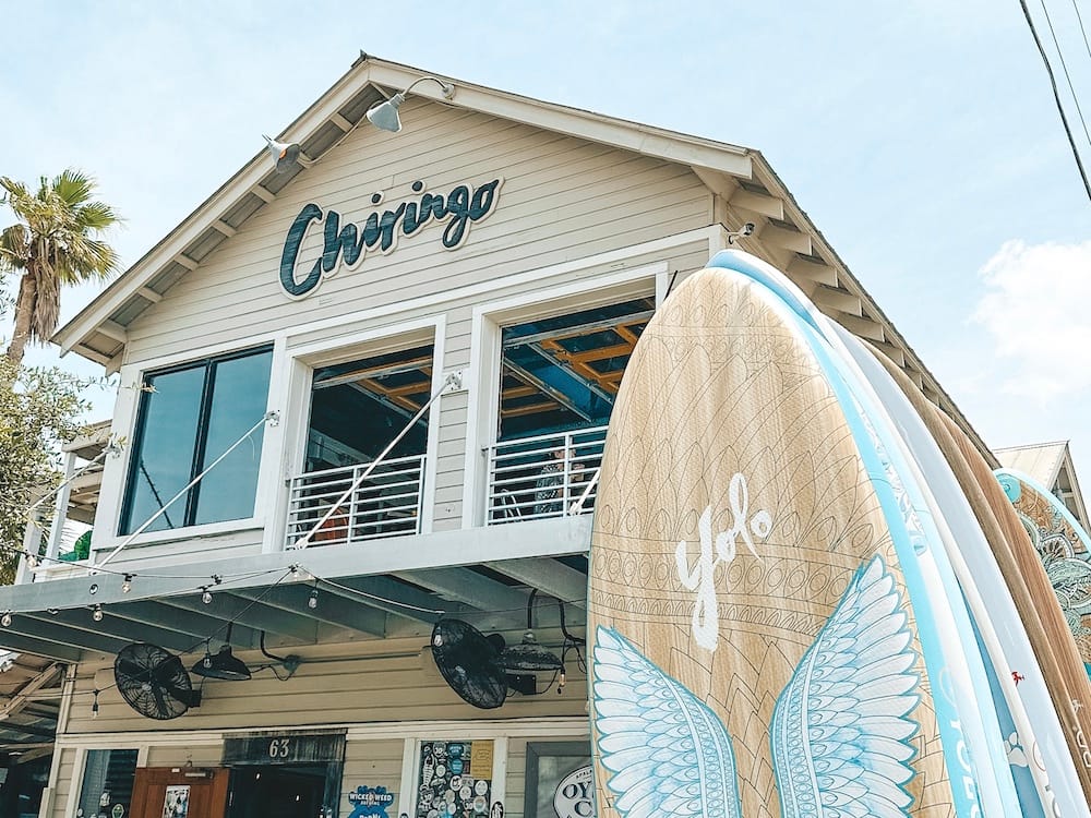 A two-story bar in Grayton Beach called Chiringo, with tan horizontal siding and surfboards out front that say "YOLO" and have blue wings.