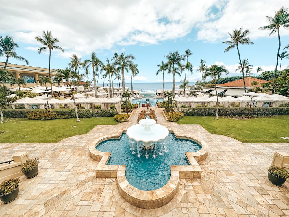 A plaza with a fountain and several cabanas at a pool overlooking several palm trees and the beach at one of the best luxury resorts in Maui