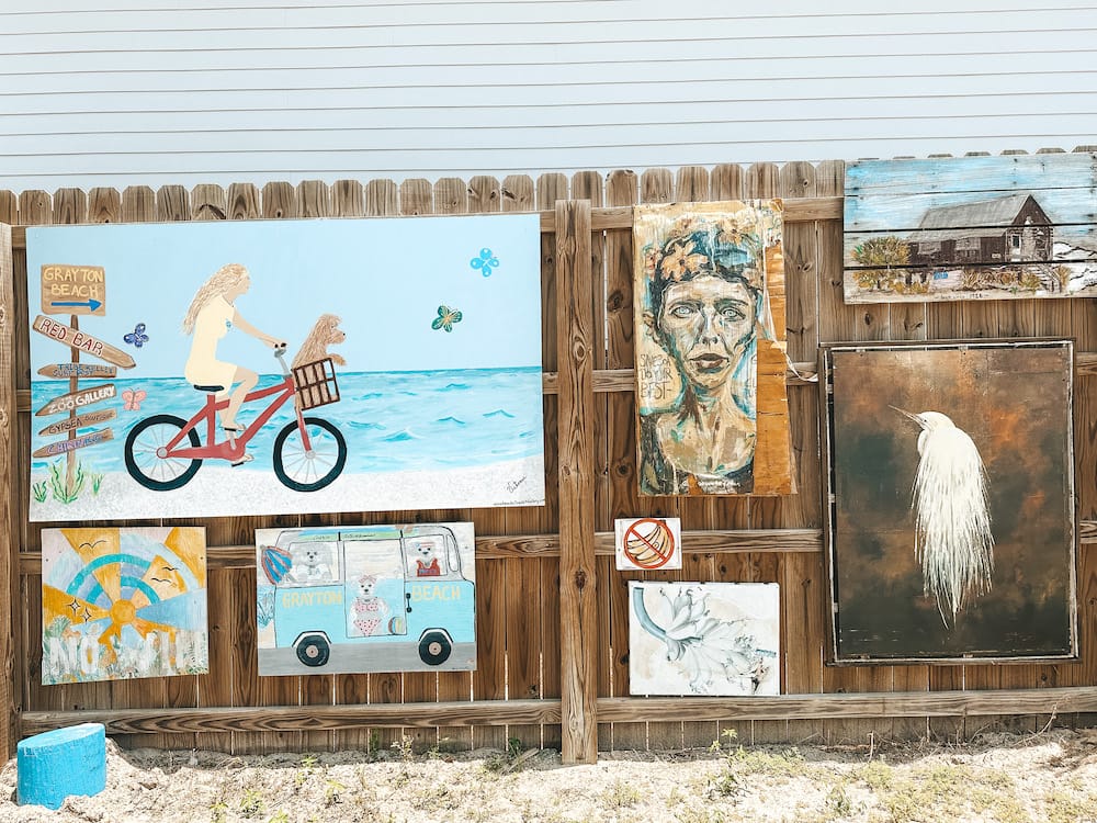 A wall of eclectic artwork In Grayton Beach, including a white bird, a cabin, a girl on a bike with a dog in the basket biking along the beach, and more.