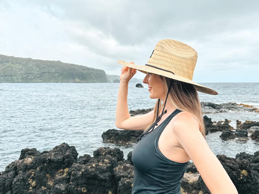 A woman standing in a black tank top and straw hat in front of the black rocky overlook at the Ke'Anae Lookout, one of the best stops on a Road to Hana itinerary