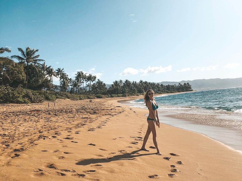 A girl in a green swimsuit standing on a beach on Oahu's North Shore with golden sand, green palm trees, and blue waves lapping the shore.