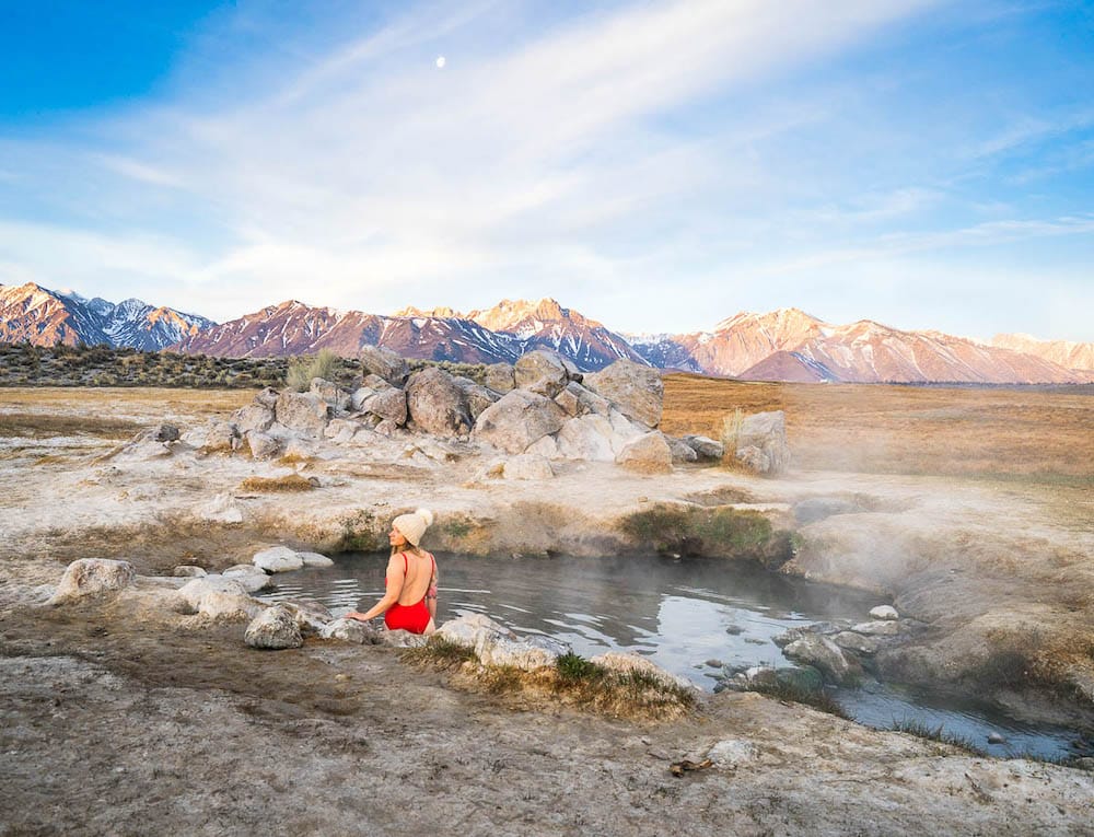 A woman in a red swimsuit and beanie sitting in a hot spring with snow-covered mountains in the background.