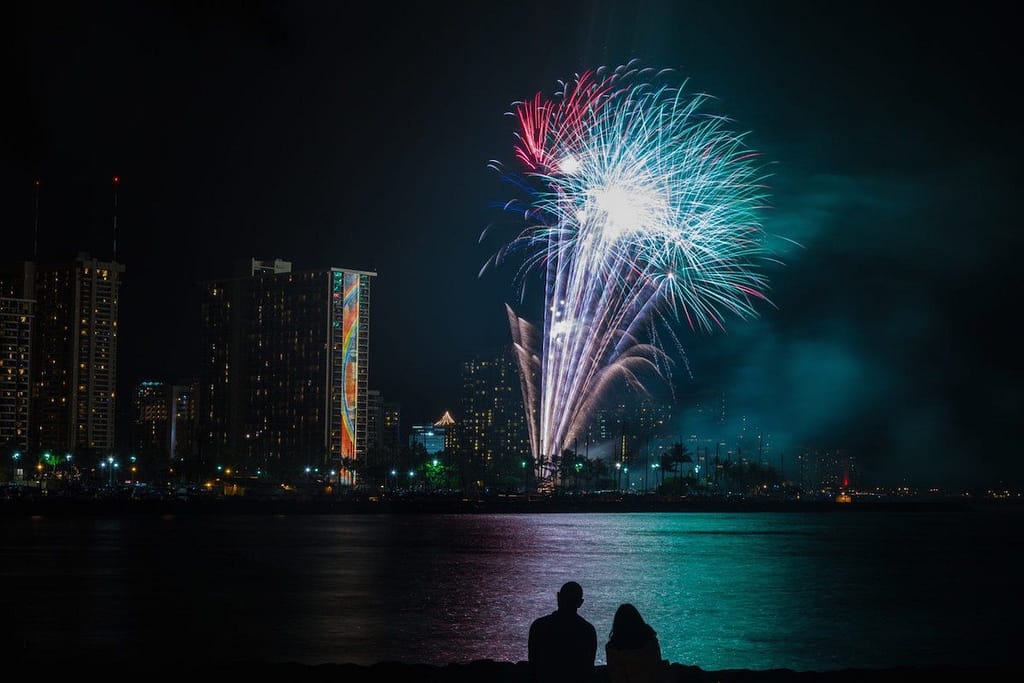 A fireworks display lighting up the night sky in Waikiki right over the Pacific Ocean with two people in the foreground.