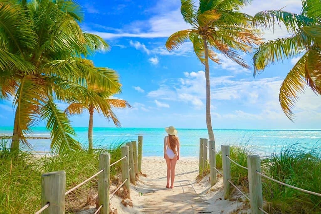 A woman in a hat, white bathing suit, and long red hair walking on a pathway to a beach surrounded by palm trees and with gorgeous blue water in the background
