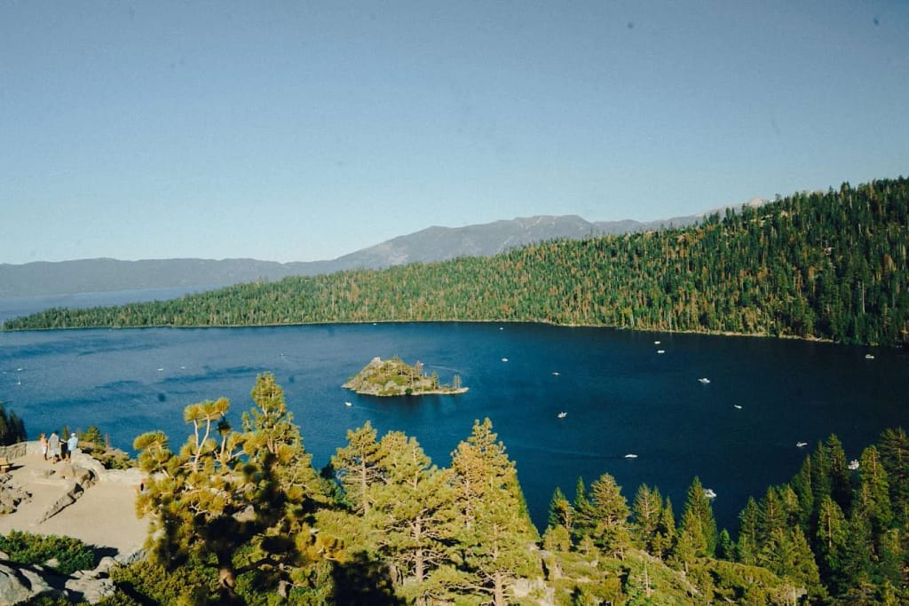 A deep blue lake with several green pine trees and granite rocks surrounding it with a small green island in the center