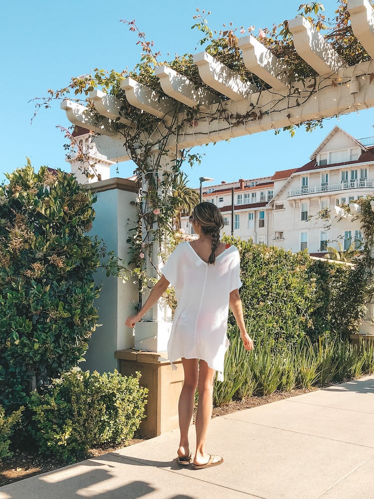 La Jolla or Coronado: A girl walking under a white archway with vines around it with a historic hotel in the background