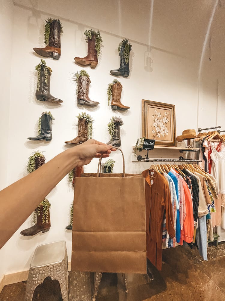 A brown paper shopping bag being held up in front of a clothes rack and cowboy boots hanging on the wall with plants growing from them in The Find at The Factory at Franklin