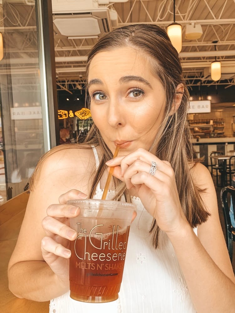 A woman with brown hair in a white dress drinking an iced tea from a straw.
