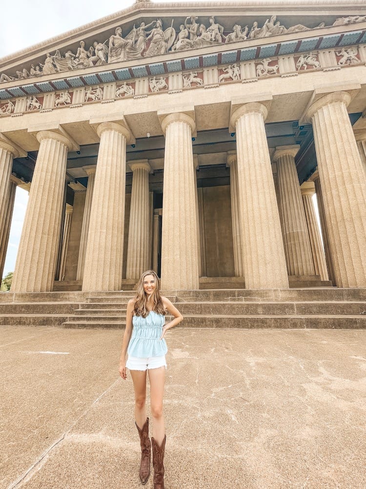 A woman wearing cowboy boots, white shorts, and a blue tank top standing in front of The Parthenon in Centennial Park Nashville