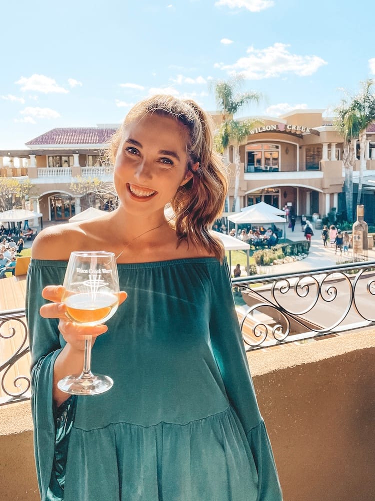 A woman standing in front of Wilson Creek Winery with a glass of white wine wearing a green off-the-shoulderr shirt on one of the best wine tours in Temecula.