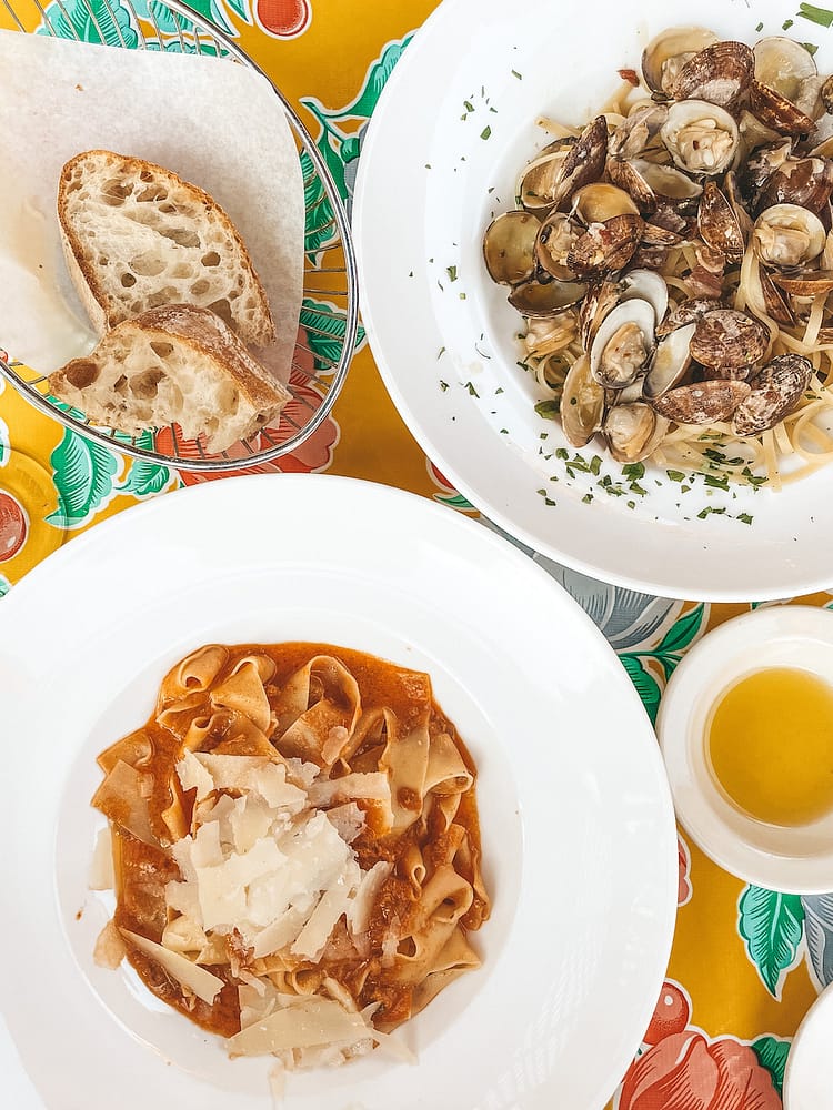Two pasta dishes, bread, and olive oil sitting on a table with a yellow tablecloth with colorful flowers on it.