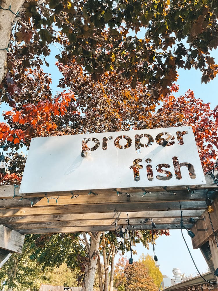 A sign that says Proper Fish in front of a tree with red leaves.