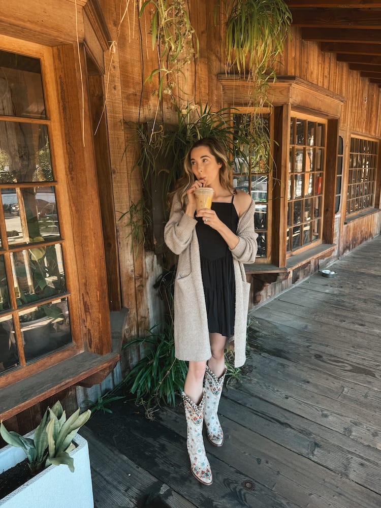 A woman holding an iced coffee on a wooden porch wearing a black dress, tan sweater, and white cowboy boots