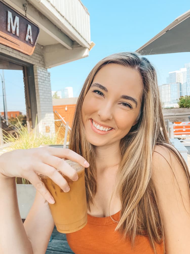 A woman with long brown hair in a rust orange tank top smiling while holding an iced coffee