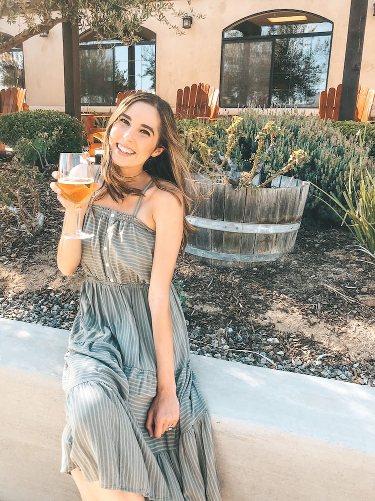 A woman sitting on a stucco half wall holding a glass of white wine in a green dress sitting in front of plants, cacti, and more.