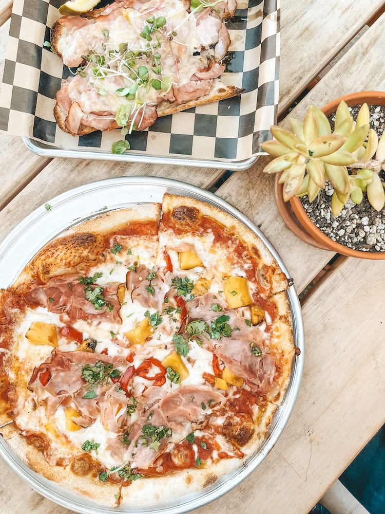 A pizza with pineapple, ham, and other toppings and a open-faced sandwich sitting on a table.