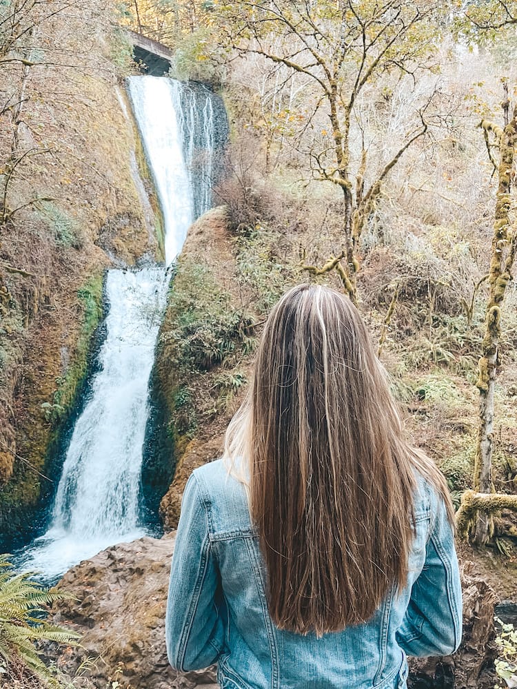 A woman with brown hair and a denim jacket standing in front of a waterfall