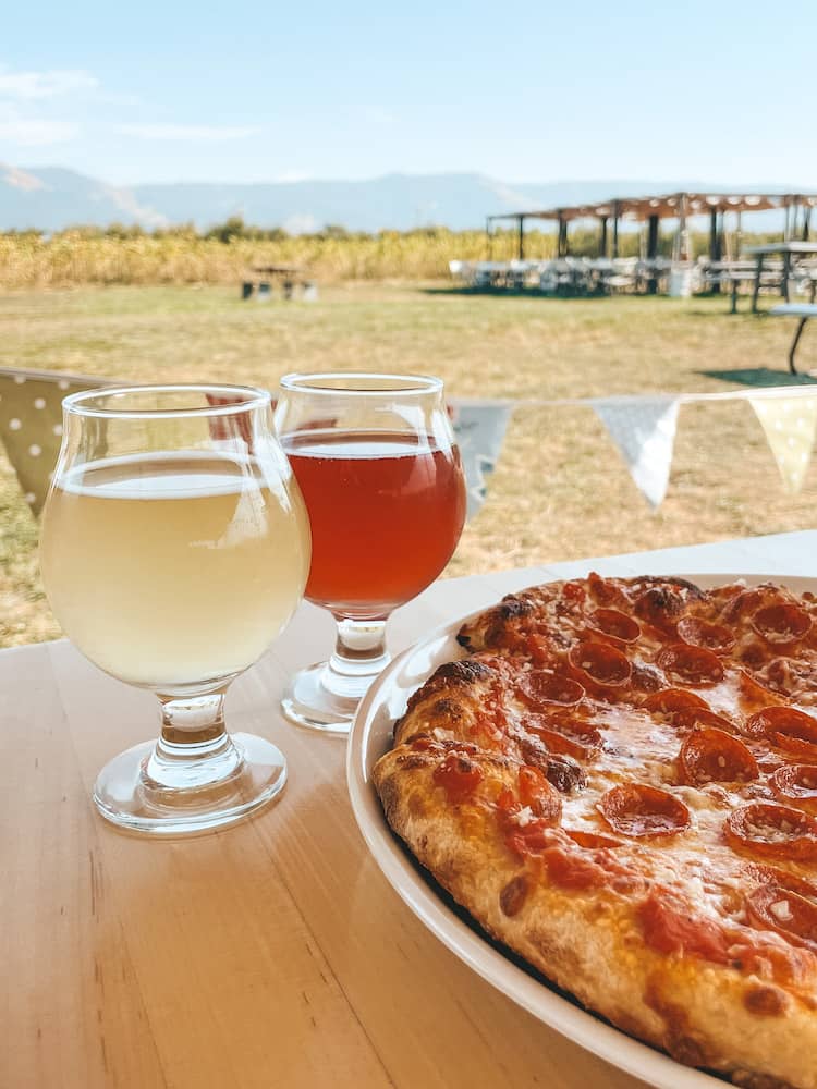 A red and yellow cider and a pizza sitting on a wooden table with beautiful scenery in the background.