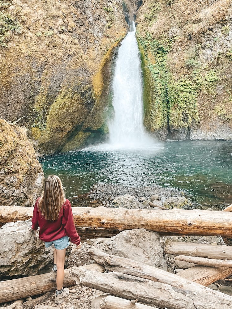 A woman in a red sweatshirt and hiking boots standing in front of a waterfall cascading into a blue pool.