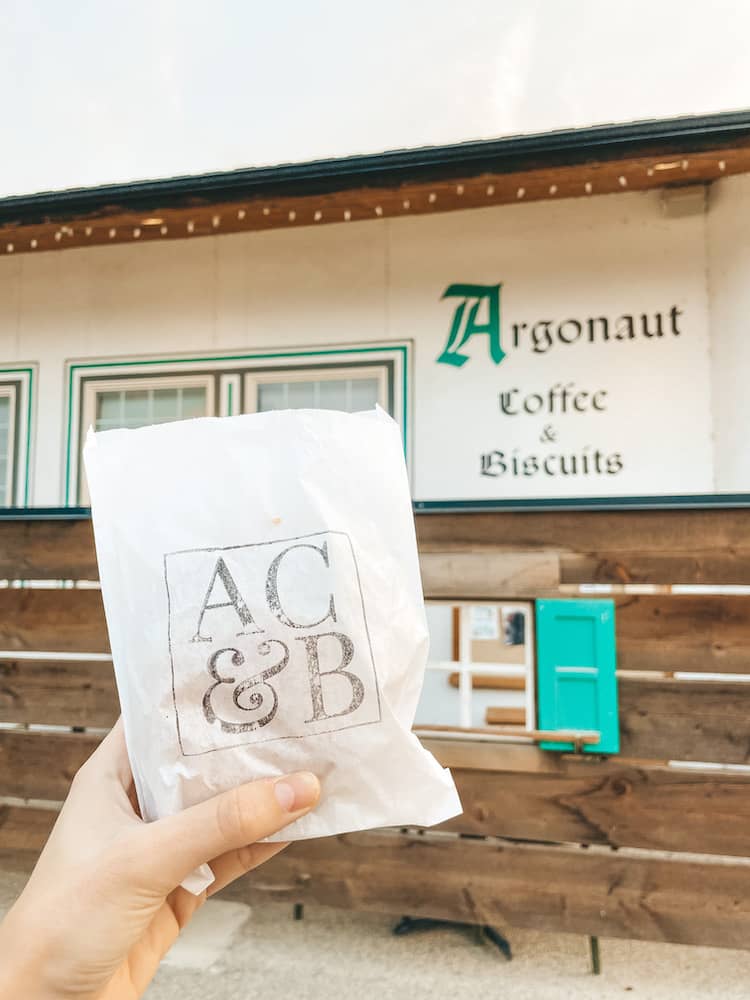 A hand holding a bag with a sandwich inside in front of a sign that says Argonaut Coffee & Biscuits