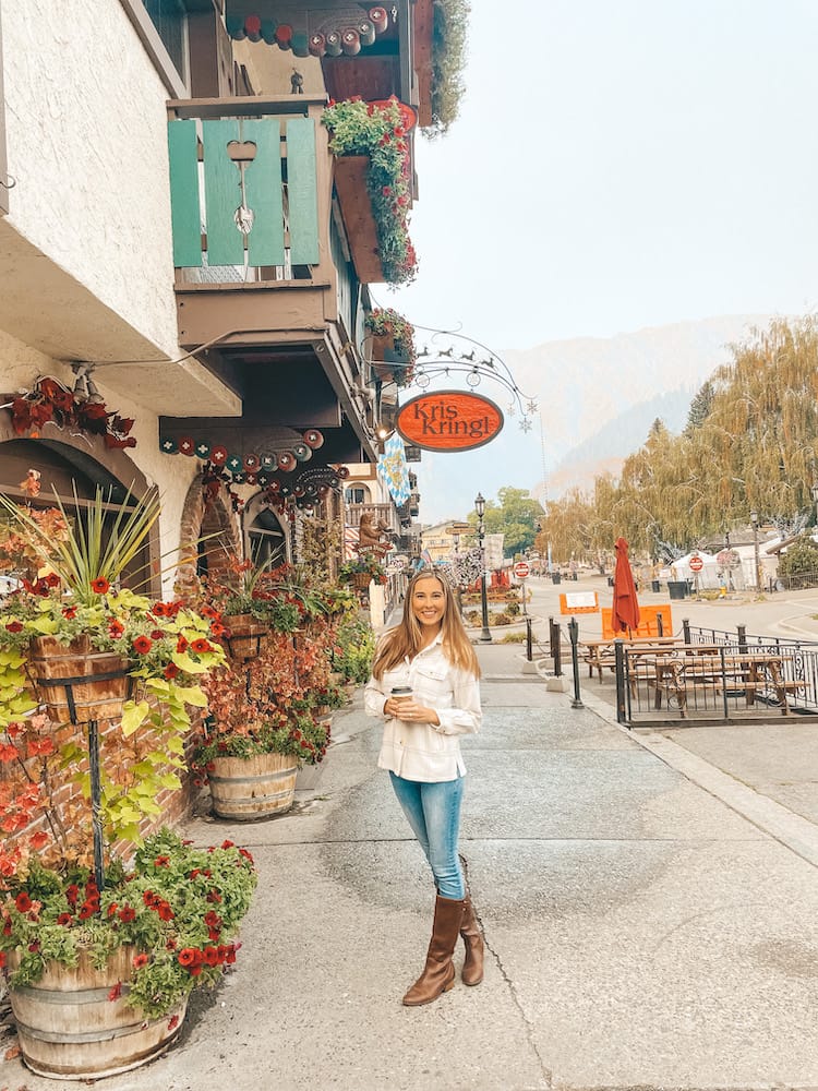 A woman in a white flannel, jeans, and brown boots holding a cup of coffee and standing in Downtown Leavenworth.