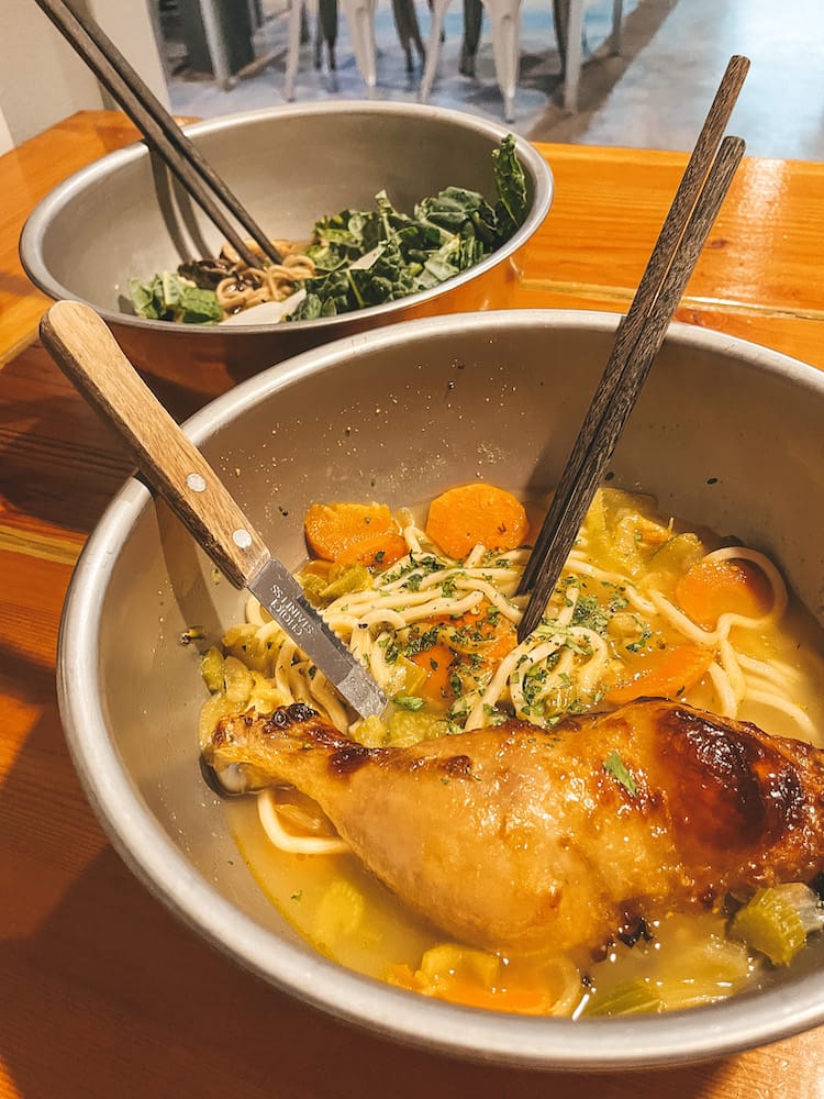 Two bowls of noodles and broth and one with a giant chicken leg inside.