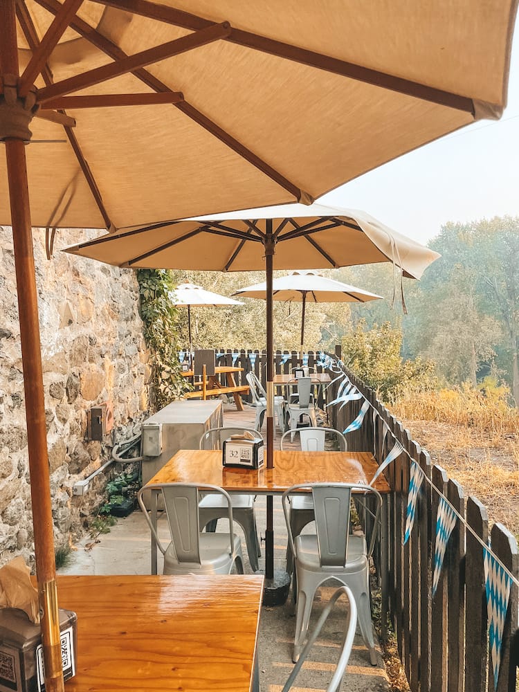 An outdoor restaurant patio with large tan umbrellas and tables overlooking a forested area.