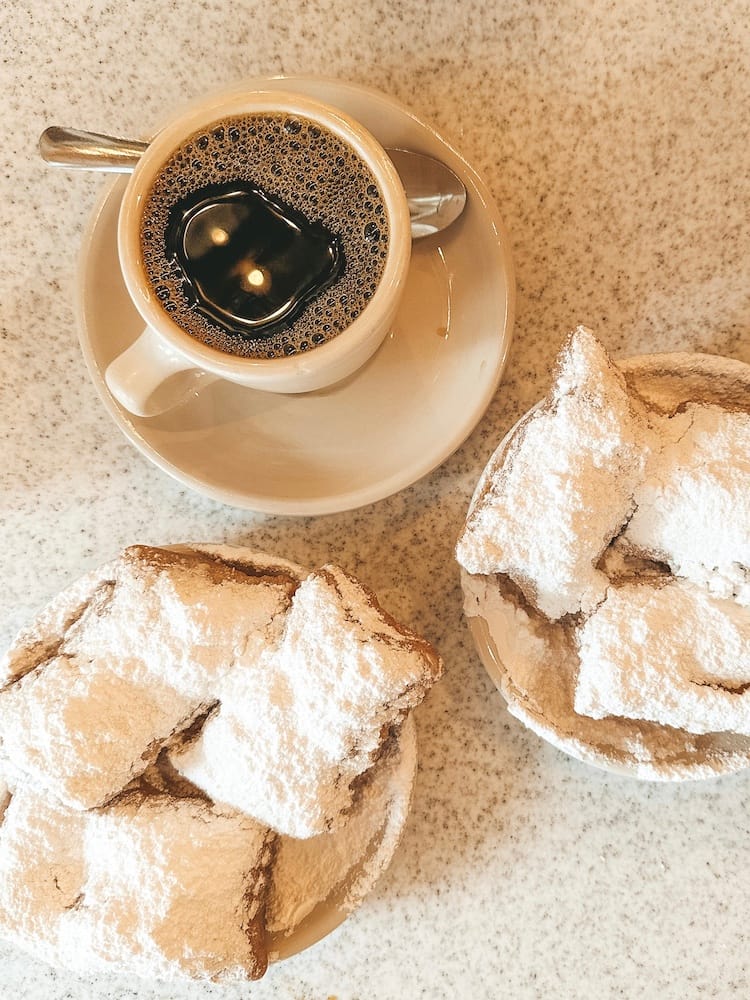 Two plates of beignets and a black coffee sitting on white plates.