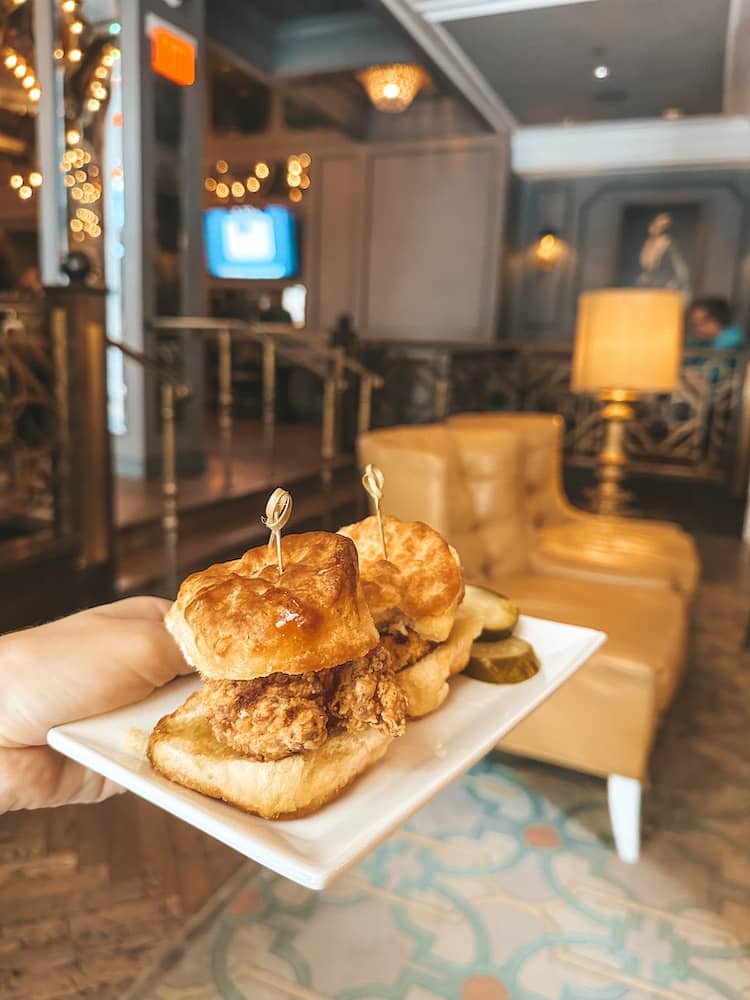 Two fried chicken biscuit sandwiches on a white rectangular plate being held in front of a lounge in New Orleans.