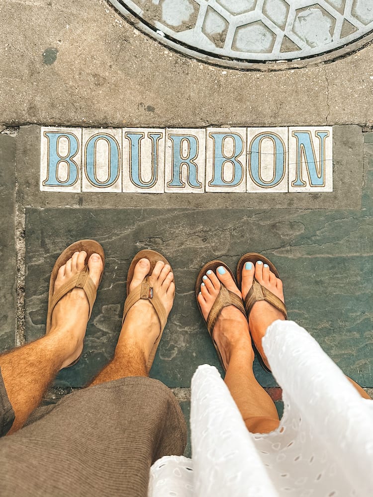 A man and a women standing in flip flops on a sidewalk with the letters "Bourbon" in the concrete with tile lettering.