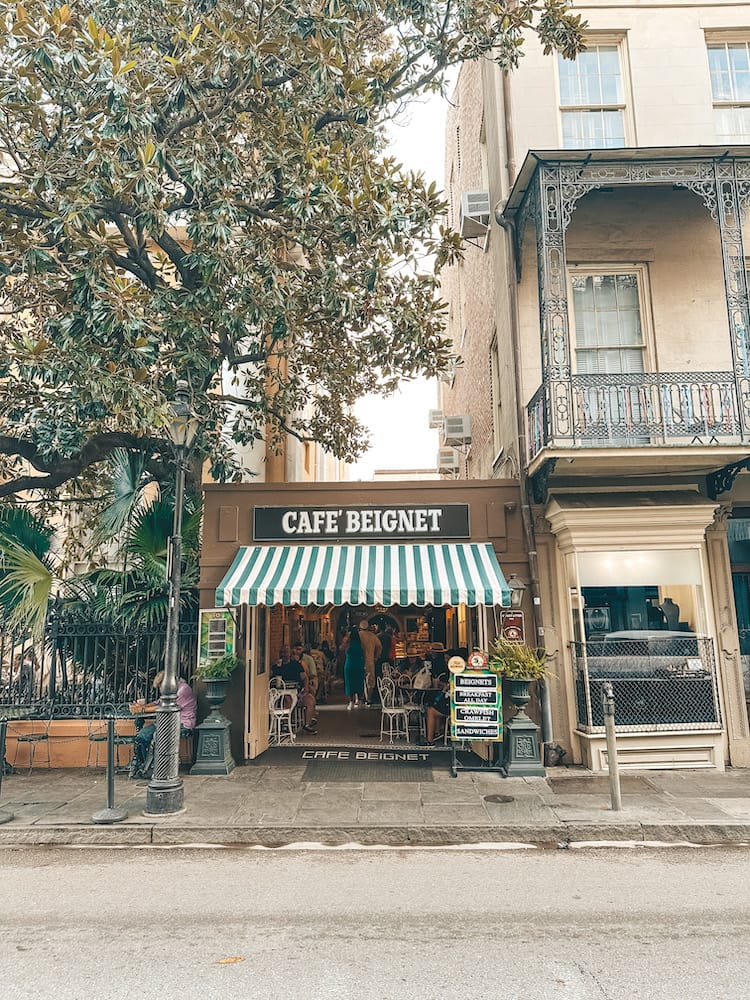 The exterior of Cafe Beignet in New Orleans with a white and green striped shade and trees surrounding the building.