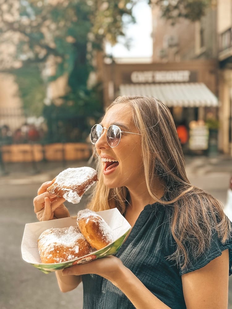 A woman in a black dress and sunglasses is holding a carton of beignets and about to put one in her mouth.