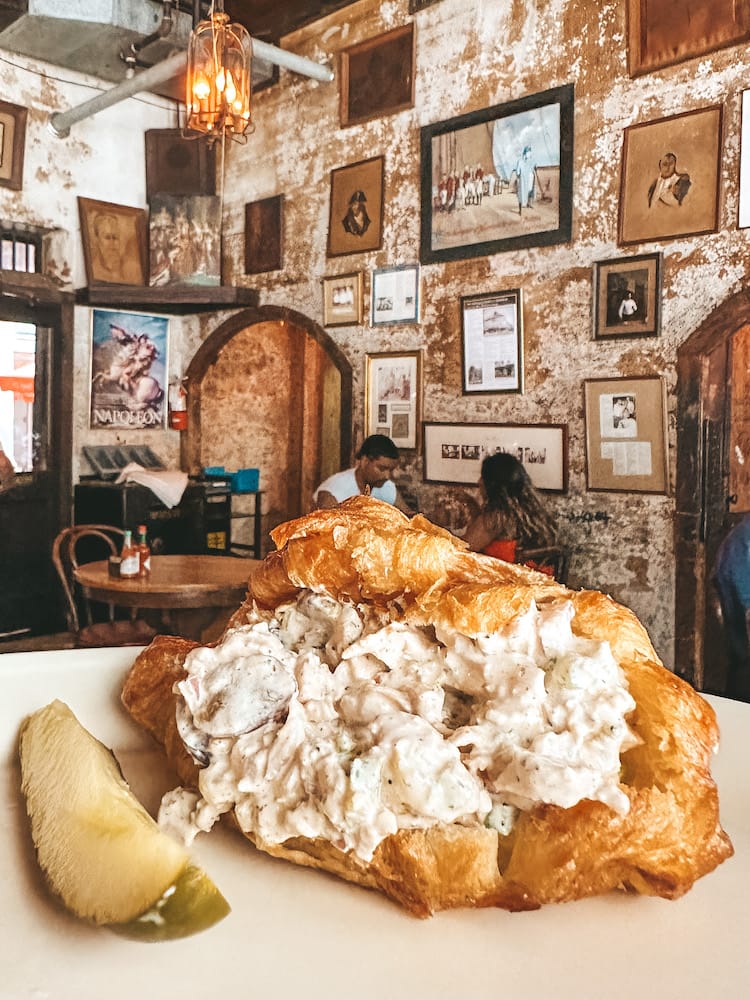 A chicken salad sandwich on a croissant in front of an old wall with lots of historic paintings.