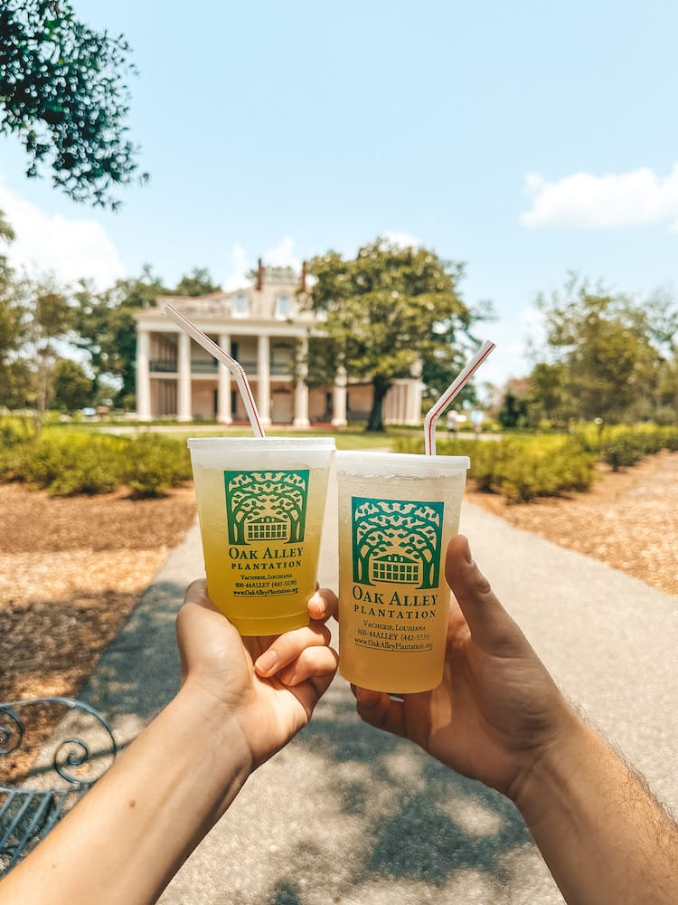 Two hands holding iced drinks in front of the big house on Oak Alley Plantation.
