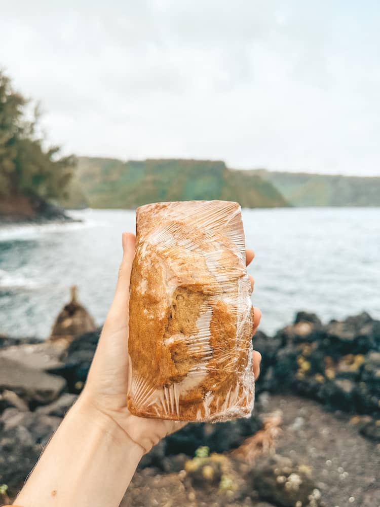 A loaf of banana bread wrapped in saran wrap being held in front of an ocean in Maui on the Road to Hana